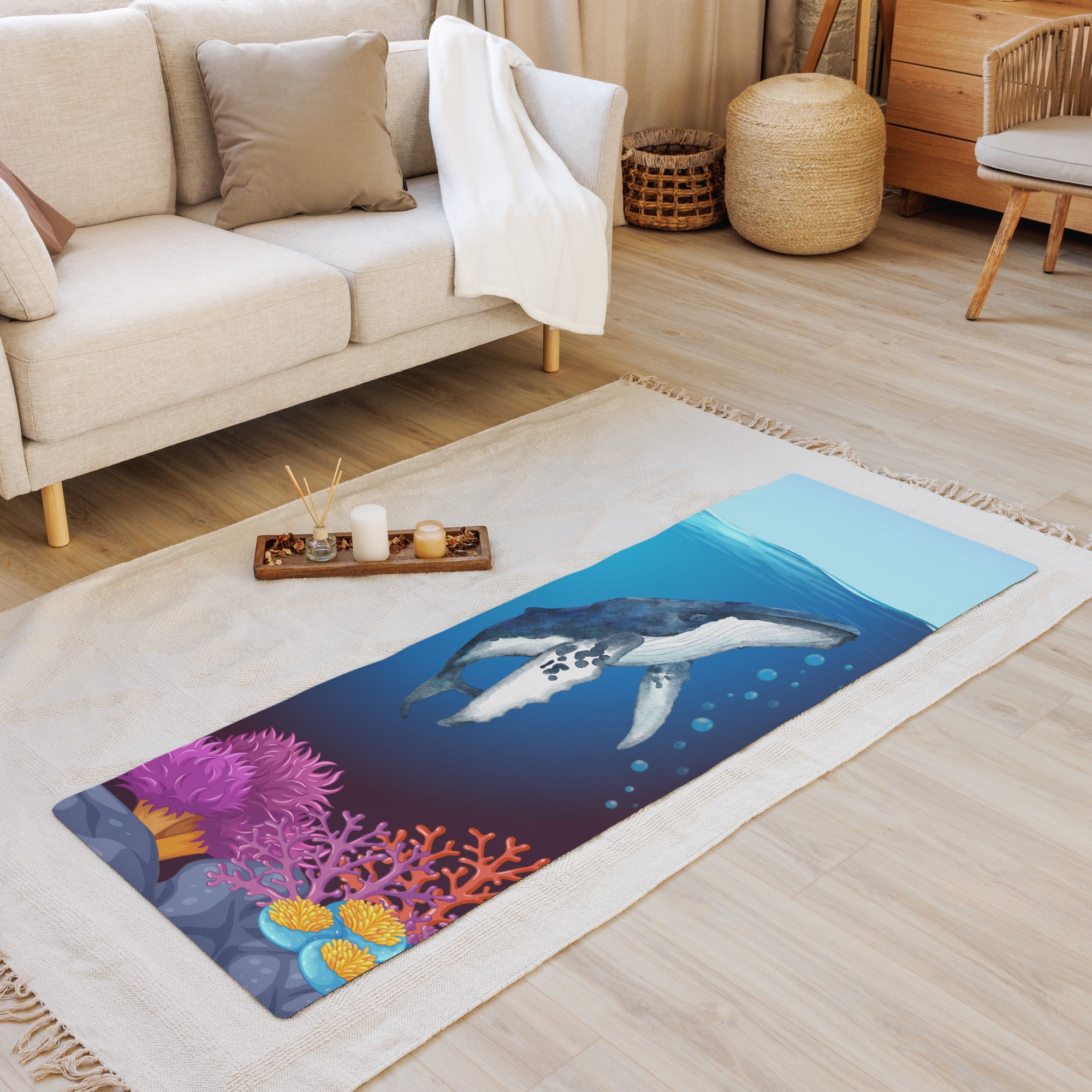 A colorful yoga mat featuring an underwater scene with a humpback whale, placed on a wooden floor next to a couch and a small wooden stool with a candle holder.