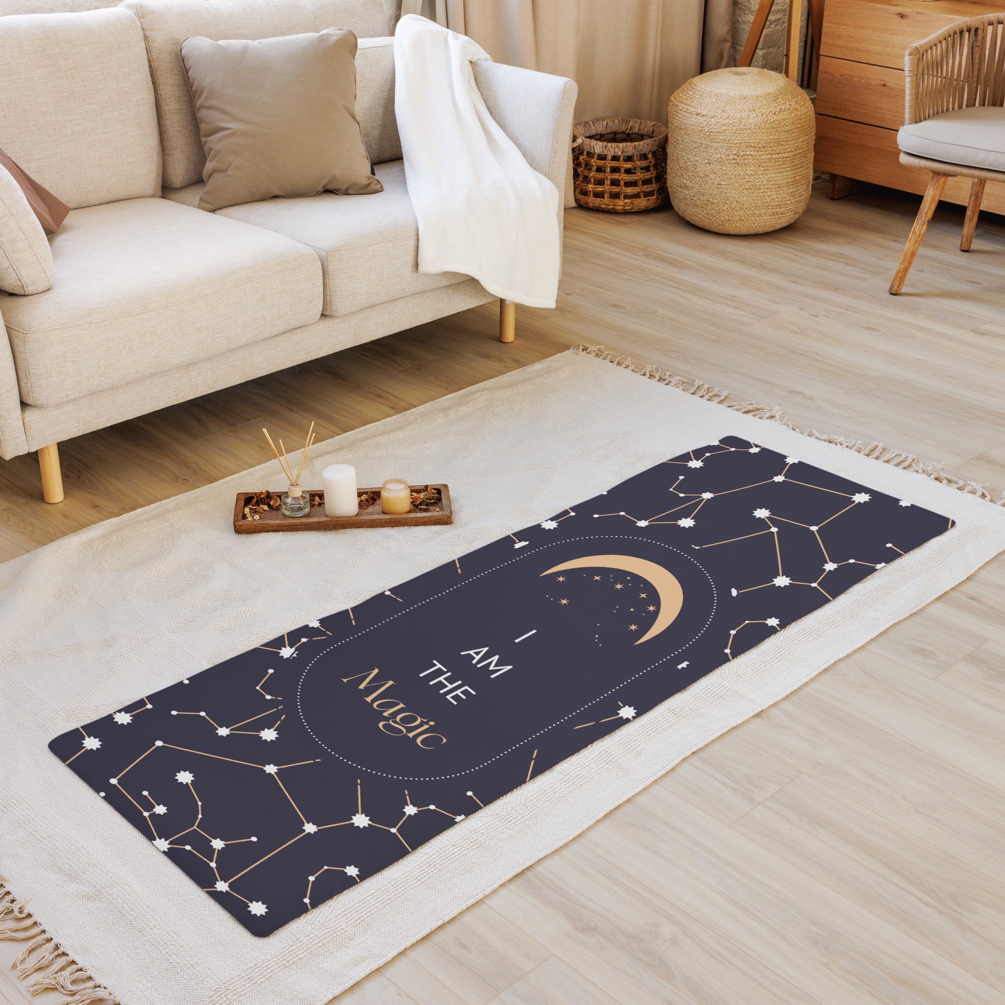 A yoga mat with a black and gray design featuring stars and the text 'I AM THE MAGIC'. The mat is placed on a wooden floor in a home setting, with a couch, chair, and candle holder in the background.