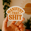 Hand holding a sticker with 'Manifest That Shit' text against a blurred indoor background