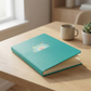 Turquoise notebook with a rainbow design on a wooden table with a cup and plant in the background.