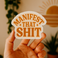 Hand holding a sticker with 'Manifest That Shit' text against a blurred indoor background