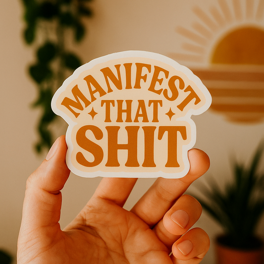 Hand holding a sticker with 'Manifest That Shit' text against a blurred indoor background