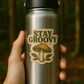 Hand holding a silver water bottle with 'Stay Groovy' and mushroom design against a blurred forest background