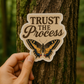 Hand holding a 'Trust the Process' sticker with a butterfly design against a tree background