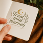 Hand holding a small notebook with a 'Trust your journey' sticker on a textured surface.